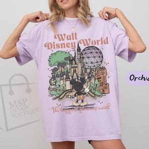 May include: Orchid-colored t-shirt with a graphic of Walt Disney World. The design includes a castle, Spaceship Earth, and Mickey Mouse. Text reads "Walt Disney World" and "the happiest place on earth."
