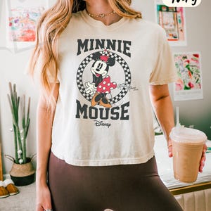 以下が含まれることがあります： アイボリー色のボックス型Tシャツ。ミニーマウスのグラフィックが白黒のチェック柄の円の中に描かれています。「MINNIE MOUSE」の文字が太字の黒でデザインされ、下にディズニーのロゴがあります。このシャツは「COMFORT COLORS」コレクションの一部です。