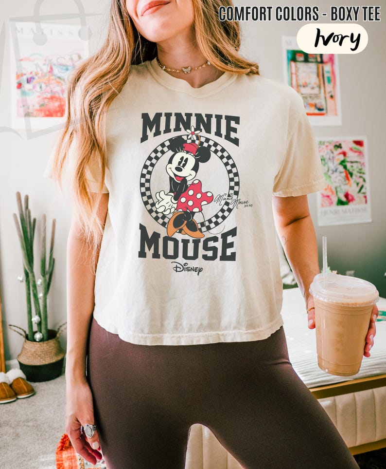 以下が含まれることがあります： アイボリー色のボックス型Tシャツ。ミニーマウスのグラフィックが白黒のチェック柄の円の中に描かれています。「MINNIE MOUSE」の文字が太字の黒でデザインされ、下にディズニーのロゴがあります。このシャツは「COMFORT COLORS」コレクションの一部です。