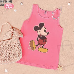 May include: Pink tank top featuring a Mickey Mouse graphic. The tank top has a round neckline and is made of a soft material. The words "COMFORT COLORS TANK TOP Water Meln" are in the top left corner. A woven handbag is in the image.