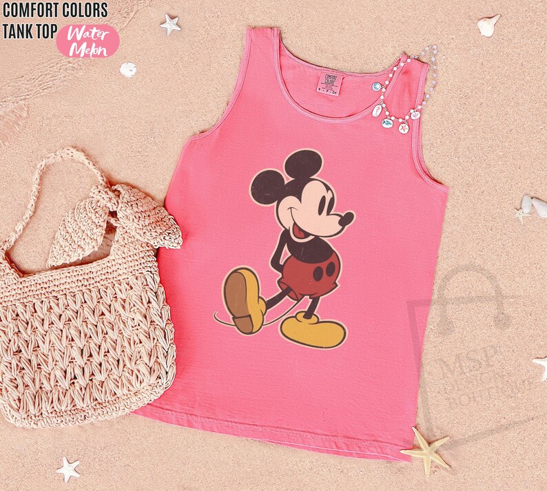 May include: Pink tank top featuring a Mickey Mouse graphic. The tank top has a round neckline and is made of a soft material. The words "COMFORT COLORS TANK TOP Water Meln" are in the top left corner. A woven handbag is in the image.