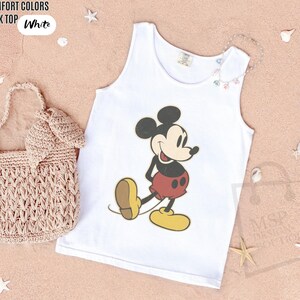 May include: White tank top featuring a classic Mickey Mouse graphic. The cartoon character is depicted in black, red, yellow, and tan. The text "COMFORT COLORS TANK TOP White" is in the upper left corner. A woven bag and decorative seashells are also in the image.