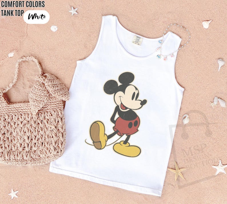 May include: White tank top featuring a classic Mickey Mouse graphic. The cartoon character is depicted in black, red, yellow, and tan. The text "COMFORT COLORS TANK TOP White" is in the upper left corner. A woven bag and decorative seashells are also in the image.