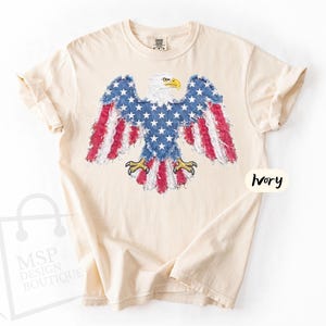 May include: An ivory short-sleeve t-shirt featuring a graphic of a bald eagle with wings spread. The eagle's body and wings are patterned with the American flag, including white stars on a blue field and red and white stripes. The text Ivory is visible.