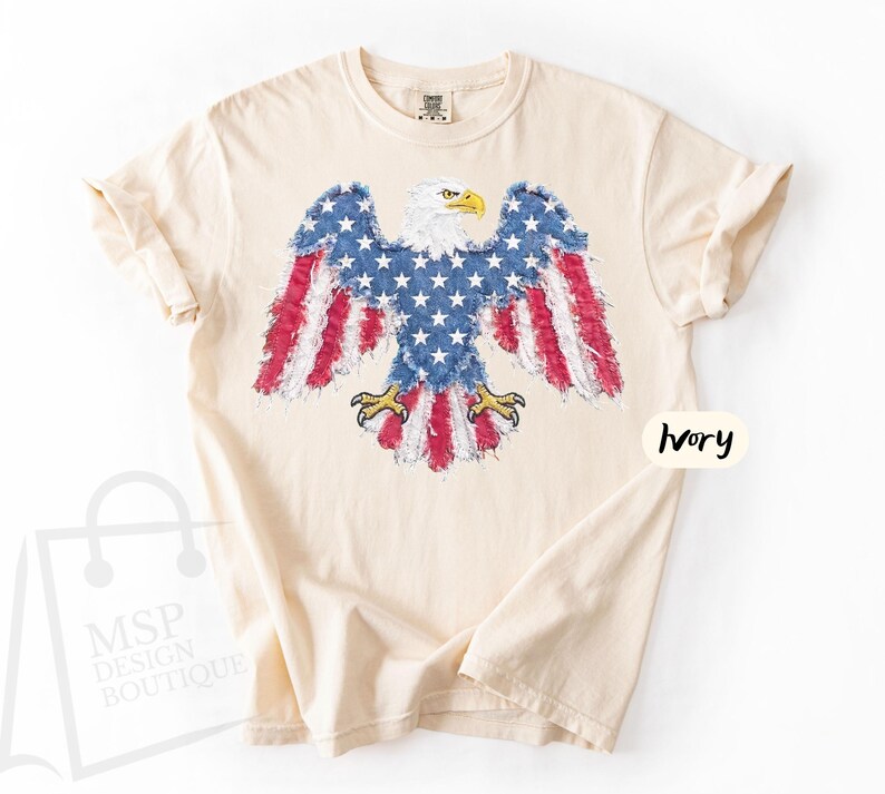 May include: An ivory short-sleeve t-shirt featuring a graphic of a bald eagle with wings spread. The eagle's body and wings are patterned with the American flag, including white stars on a blue field and red and white stripes. The text Ivory is visible.