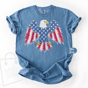 May include: A blue jean colored short-sleeve t-shirt featuring a graphic of a bald eagle with wings spread. The eagle's body is patterned with white stars on a blue background, while the wings and tail feature red and white stripes with frayed edges.