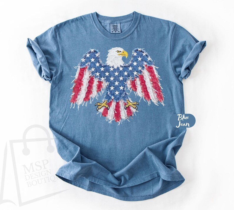 May include: A blue jean colored short-sleeve t-shirt featuring a graphic of a bald eagle with wings spread. The eagle's body is patterned with white stars on a blue background, while the wings and tail feature red and white stripes with frayed edges.