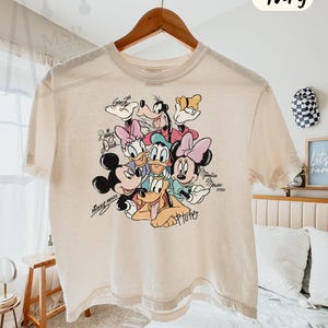 Puede incluir: Camiseta holgada de color marfil con un gr&aacute;fico colorido de personajes de Disney: Mickey y Minnie Mouse, Donald y Daisy Duck, Goofy y Pluto. La camiseta tiene mangas cortas y cuello redondo. El texto en la camiseta dice "COMFORT COLORS - BOXY TEE" e "Ivory".