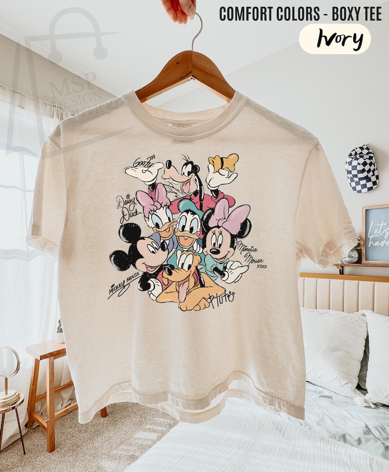 Puede incluir: Camiseta holgada de color marfil con un gr&aacute;fico colorido de personajes de Disney: Mickey y Minnie Mouse, Donald y Daisy Duck, Goofy y Pluto. La camiseta tiene mangas cortas y cuello redondo. El texto en la camiseta dice "COMFORT COLORS - BOXY TEE" e "Ivory".