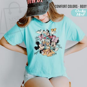Puede incluir: Una camiseta holgada de color azul claro con un gr&aacute;fico de personajes de Disney, incluyendo Mickey Mouse, Minnie Mouse, Donald Duck, Daisy Duck, Goofy y Pluto. La camiseta tiene el texto "COMFORT COLORS - BOXY TEE" y "Chalky Mint".