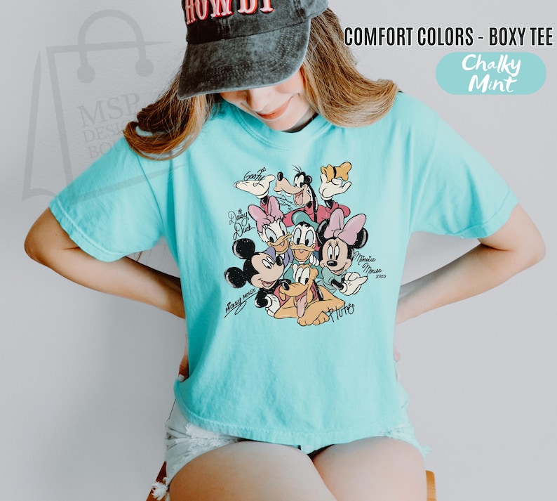Puede incluir: Una camiseta holgada de color azul claro con un gr&aacute;fico de personajes de Disney, incluyendo Mickey Mouse, Minnie Mouse, Donald Duck, Daisy Duck, Goofy y Pluto. La camiseta tiene el texto "COMFORT COLORS - BOXY TEE" y "Chalky Mint".