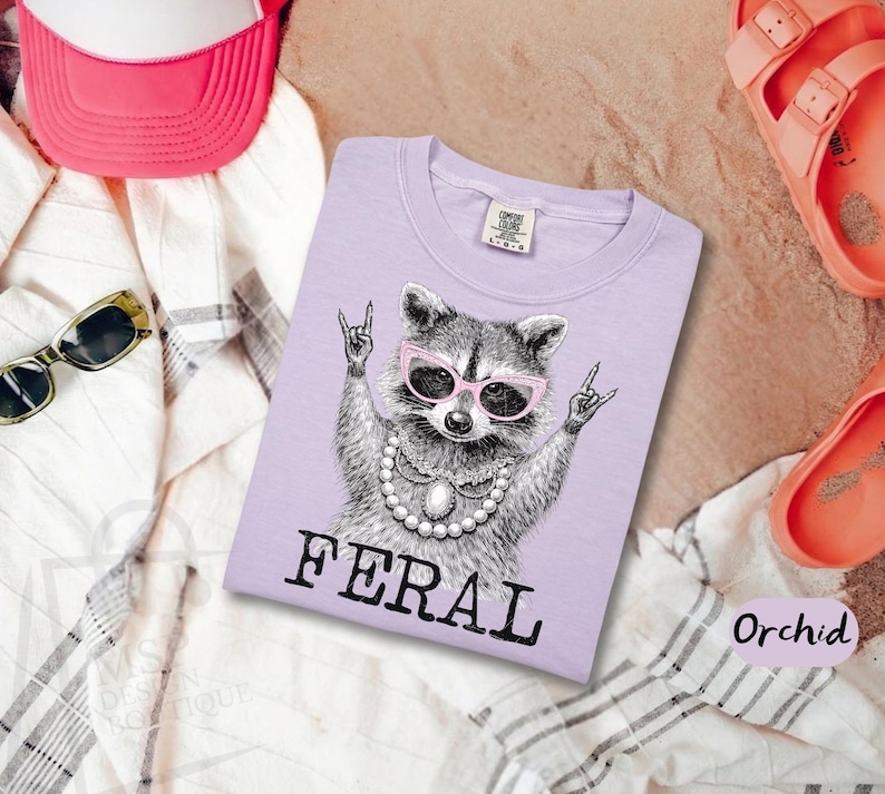 May include: A light purple t-shirt with a graphic of a raccoon wearing pink sunglasses and a pearl necklace, making a rock and roll hand gesture. The word "FERAL" is printed in black block letters below the raccoon.
