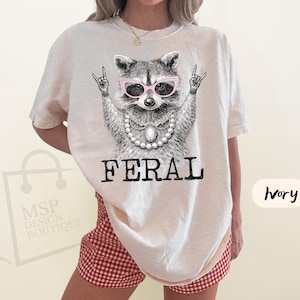 May include: Ivory-colored t-shirt featuring a raccoon wearing pink glasses and a pearl necklace, with the word "FERAL" in bold black letters. The raccoon is making a rock and roll hand gesture. The shirt is a relaxed fit.