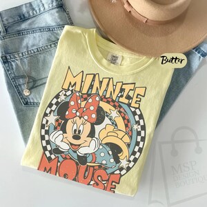 May include: A yellow t-shirt featuring a graphic of Minnie Mouse with a red polka dot bow. The shirt has the words "MINNIE MOUSE" in a retro font. The shirt is paired with light blue jeans and a tan hat.