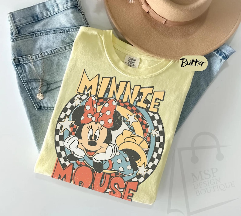 May include: A yellow t-shirt featuring a graphic of Minnie Mouse with a red polka dot bow. The shirt has the words "MINNIE MOUSE" in a retro font. The shirt is paired with light blue jeans and a tan hat.