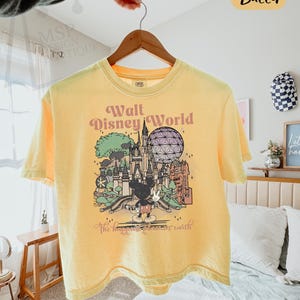 May include: Yellow Comfort Colors boxy tee featuring a graphic of Walt Disney World with Mickey Mouse. The text on the shirt reads "Walt Disney World" and "The happiest place on earth".