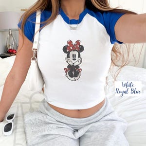 May include: White and royal blue raglan crop top featuring a Minnie Mouse graphic. The Minnie Mouse illustration has a red bow with white polka dots. The text "White Royal Blue" is in the bottom right corner.