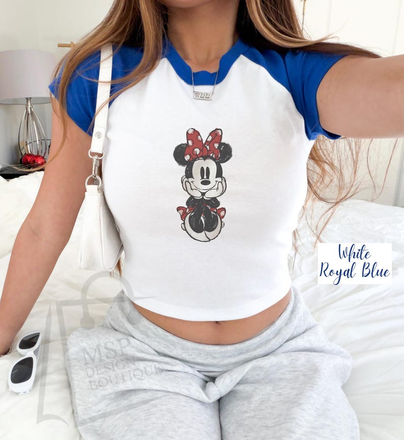 May include: White and royal blue raglan crop top featuring a Minnie Mouse graphic. The Minnie Mouse illustration has a red bow with white polka dots. The text "White Royal Blue" is in the bottom right corner.