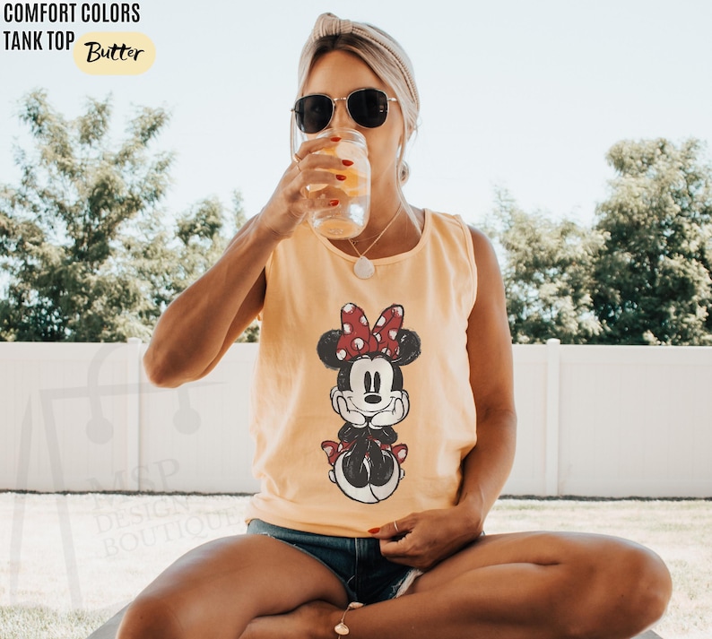 May include: A butter yellow tank top featuring a graphic print of Minnie Mouse with a red and white polka dot bow. The text COMFORT COLORS TANK TOP Butter is visible in the upper left corner, and the logo MSP DESIGN BOUTIQUE is in the lower left.