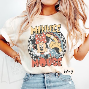May include: Ivory-colored t-shirt featuring a retro graphic of Minnie Mouse. The design includes Minnie's face, a red polka-dot bow, and the words "MINNIE MOUSE" in a vintage font. The shirt has short sleeves and a crew neck.