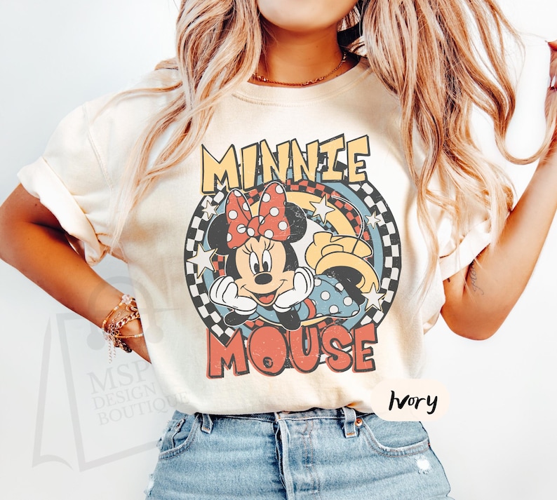 May include: Ivory-colored t-shirt featuring a retro graphic of Minnie Mouse. The design includes Minnie's face, a red polka-dot bow, and the words "MINNIE MOUSE" in a vintage font. The shirt has short sleeves and a crew neck.