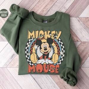 Puede incluir: Una sudadera verde militar con un gr&aacute;fico retro de Mickey Mouse. El dise&ntilde;o incluye las palabras "Mickey Mouse" en una fuente vintage, con la imagen de Mickey en el centro. La sudadera tiene mangas largas y cuello redondo.