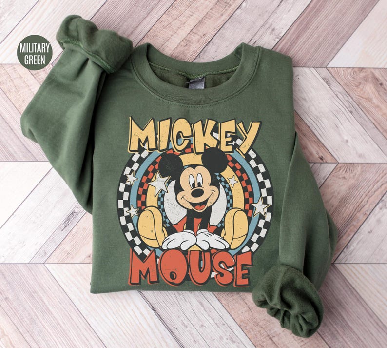 Puede incluir: Una sudadera verde militar con un gr&aacute;fico retro de Mickey Mouse. El dise&ntilde;o incluye las palabras "Mickey Mouse" en una fuente vintage, con la imagen de Mickey en el centro. La sudadera tiene mangas largas y cuello redondo.