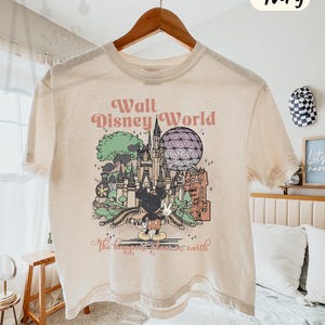 May include: Ivory-colored boxy tee featuring a graphic of Walt Disney World with Mickey Mouse. The design includes the text "Walt Disney World" and "The happiest place on earth". The shirt is hanging on a wooden hanger.
