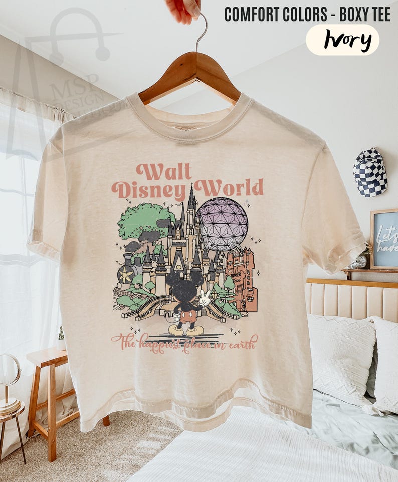 May include: Ivory-colored boxy tee featuring a graphic of Walt Disney World with Mickey Mouse. The design includes the text "Walt Disney World" and "The happiest place on earth". The shirt is hanging on a wooden hanger.