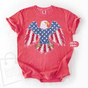 May include: A coral red short-sleeve t-shirt featuring a central graphic of a bald eagle with wings spread. The eagle is designed with a blue field of white stars on its body and wings, with red and white striped frayed fabric edges.