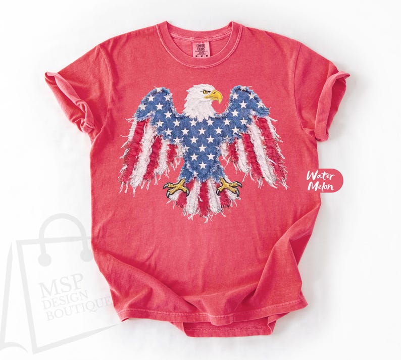 May include: A coral red short-sleeve t-shirt featuring a central graphic of a bald eagle with wings spread. The eagle is designed with a blue field of white stars on its body and wings, with red and white striped frayed fabric edges.