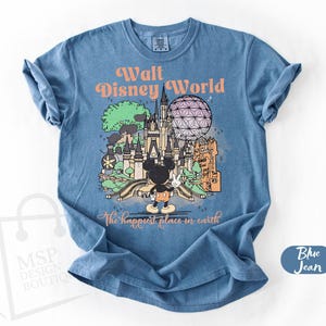 May include: Blue jean t-shirt featuring a colorful graphic of Walt Disney World with Mickey Mouse. The text reads "Walt Disney World" and "The happiest place in earth". The shirt has short sleeves and a crew neck.