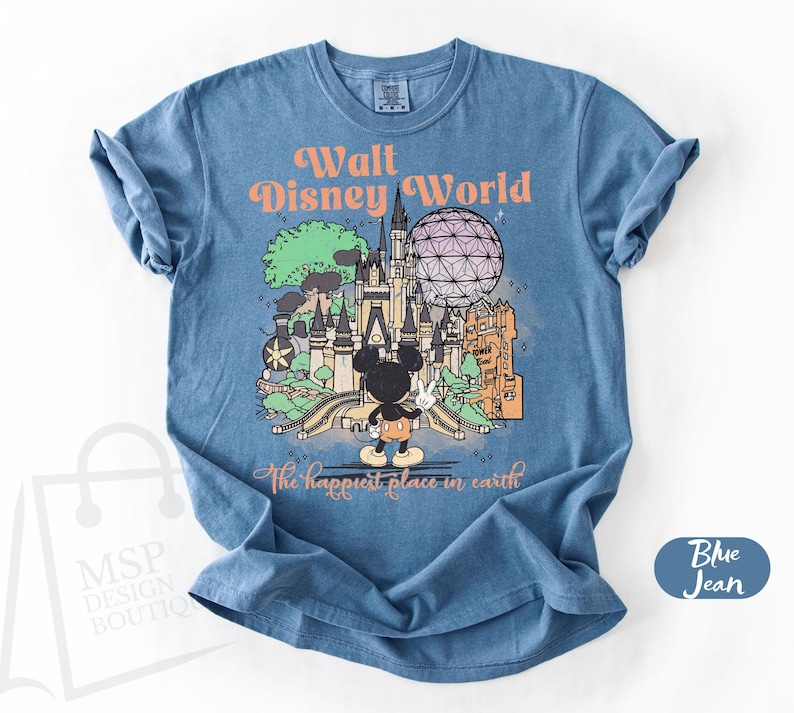 May include: Blue jean t-shirt featuring a colorful graphic of Walt Disney World with Mickey Mouse. The text reads "Walt Disney World" and "The happiest place in earth". The shirt has short sleeves and a crew neck.