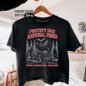 May include: A black boxy t-shirt featuring a graphic print with the text "PROTECT OUR NATIONAL PARKS" and "WHERE LEGENDS STILL ROAM FREE". The illustration depicts a forest scene with a sasquatch, a deer, an alien, a ghost, and a bat.