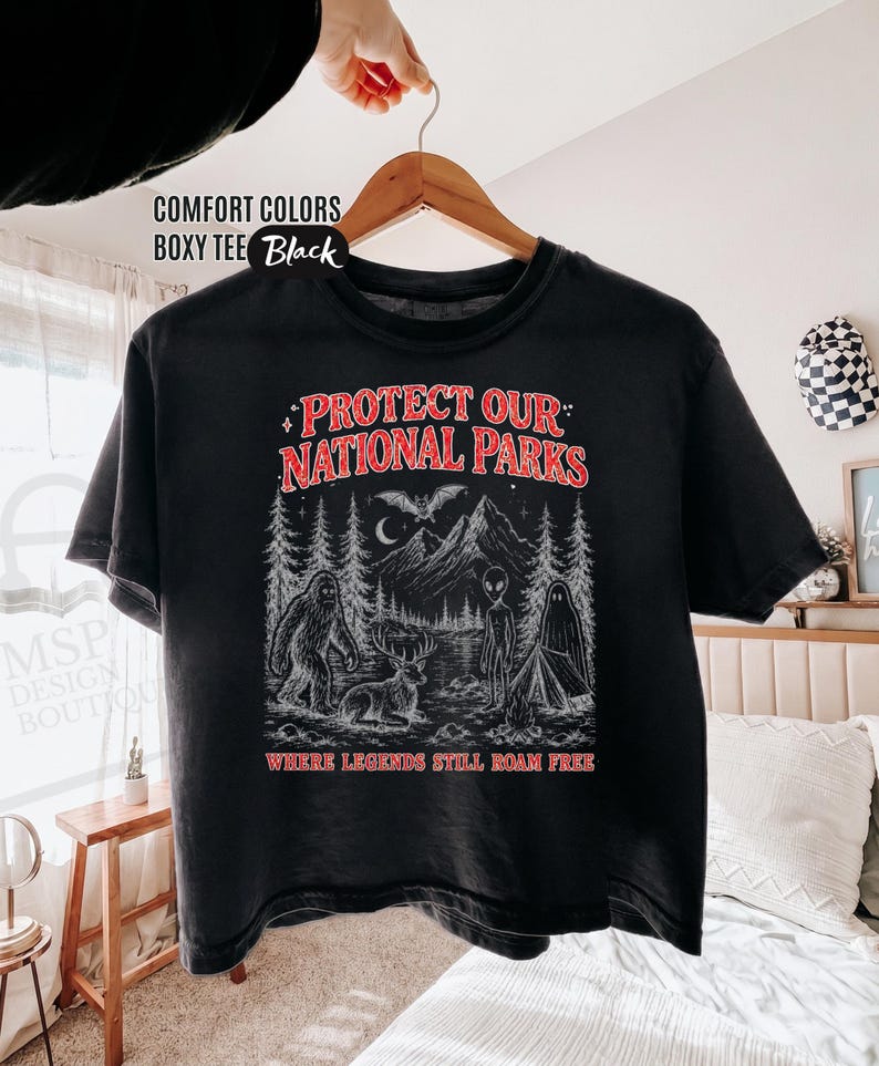 May include: A black boxy t-shirt featuring a graphic print with the text "PROTECT OUR NATIONAL PARKS" and "WHERE LEGENDS STILL ROAM FREE". The illustration depicts a forest scene with a sasquatch, a deer, an alien, a ghost, and a bat.