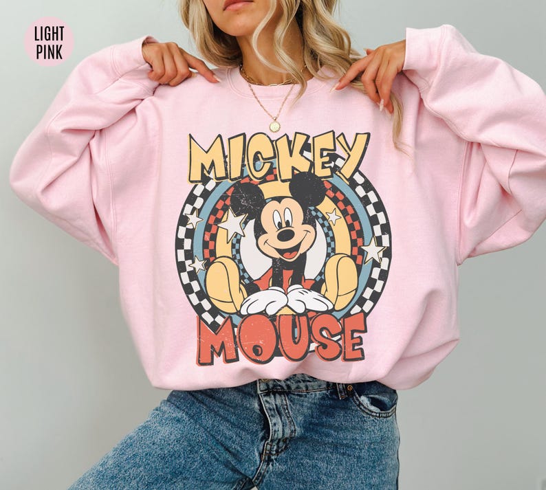 Puede incluir: Sudadera rosa claro con un gr&aacute;fico vintage de Mickey Mouse. El dise&ntilde;o incluye las palabras "Mickey Mouse" en una fuente retro, sobre un fondo circular con estrellas y un patr&oacute;n de tablero de ajedrez. La sudadera es de estilo casual.