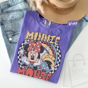 May include: A violet t-shirt featuring a graphic of Minnie Mouse with a red polka dot bow, set against a retro-style design. The shirt is paired with light blue jeans and a tan hat. The text "MINNIE MOUSE" is prominently displayed.