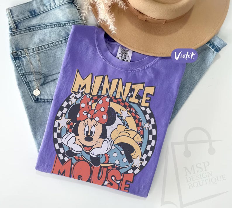 May include: A violet t-shirt featuring a graphic of Minnie Mouse with a red polka dot bow, set against a retro-style design. The shirt is paired with light blue jeans and a tan hat. The text "MINNIE MOUSE" is prominently displayed.