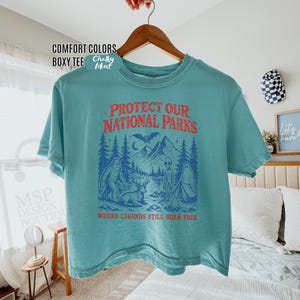 May include: A teal boxy t-shirt featuring a graphic print with the text "PROTECT OUR NATIONAL PARKS" in red and "WHERE LEGENDS STILL ROAM FREE" in red. The graphic depicts a forest, mountains, a bat, a deer, a ghost, and an alien.