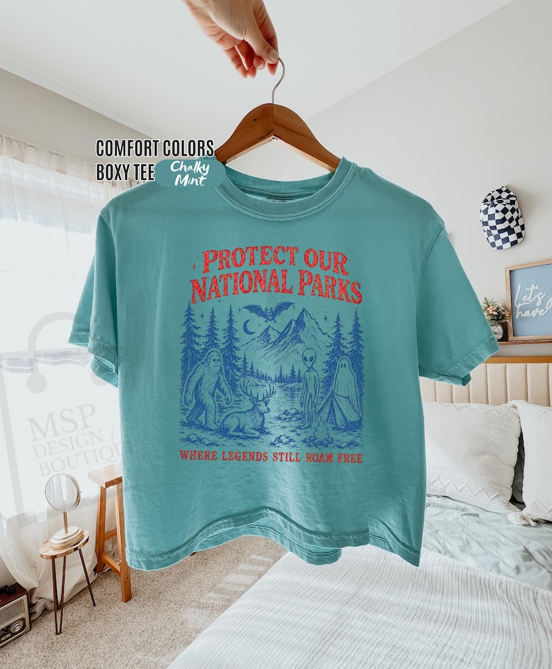 May include: A teal boxy t-shirt featuring a graphic print with the text "PROTECT OUR NATIONAL PARKS" in red and "WHERE LEGENDS STILL ROAM FREE" in red. The graphic depicts a forest, mountains, a bat, a deer, a ghost, and an alien.
