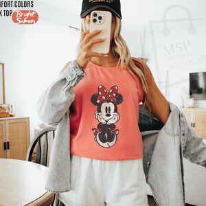 May include: A bright salmon colored tank top featuring a graphic print of Minnie Mouse with a red polka dot bow. The tank top is worn with a light blue denim button-down shirt and white shorts. Text on the image reads COMFORT COLORS TANK TOP.