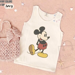 May include: Ivory tank top with a Mickey Mouse graphic. The sleeveless shirt features a classic Mickey Mouse design in black, red, yellow, and brown. A pink woven handbag is next to the tank top.
