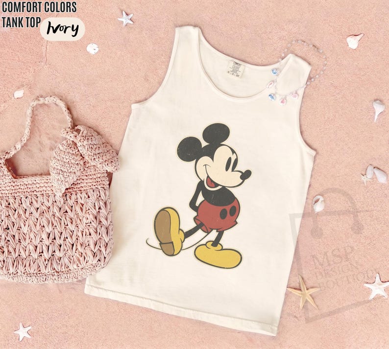 May include: Ivory tank top with a Mickey Mouse graphic. The sleeveless shirt features a classic Mickey Mouse design in black, red, yellow, and brown. A pink woven handbag is next to the tank top.