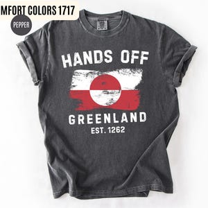 May include: Dark gray t-shirt with the words "HANDS OFF GREENLAND" in white, along with a Greenland flag design. The shirt also includes the text "EST. 1262". The shirt is made of a soft, comfortable material.