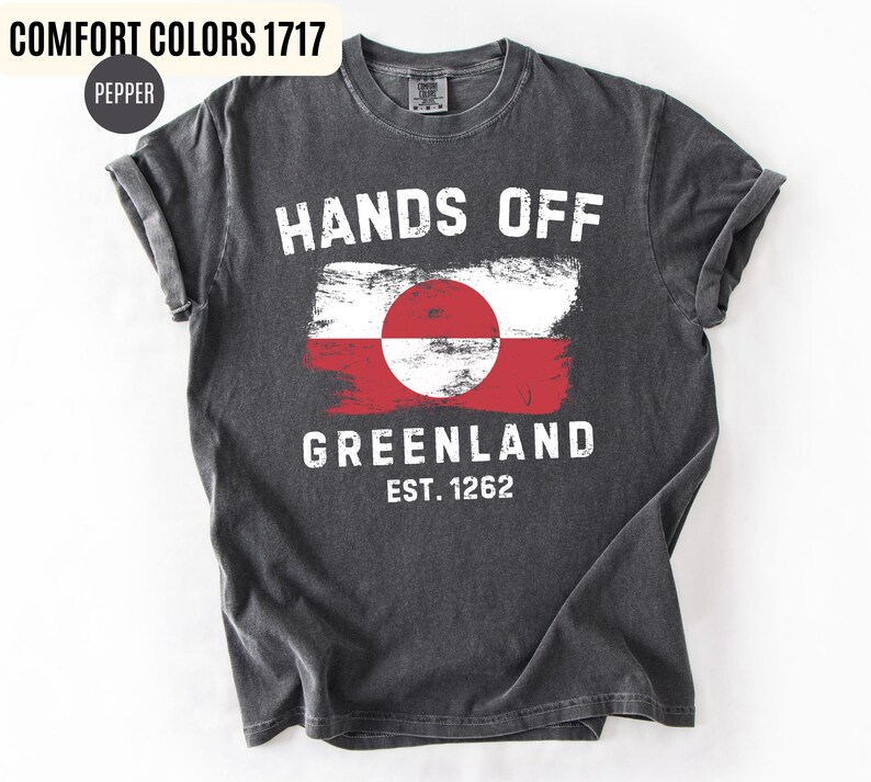 May include: Dark gray t-shirt with the words "HANDS OFF GREENLAND" in white, along with a Greenland flag design. The shirt also includes the text "EST. 1262". The shirt is made of a soft, comfortable material.