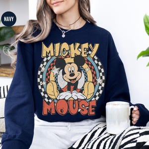 Puede incluir: Sudadera azul marino con un dise&ntilde;o de Mickey Mouse de estilo vintage. El dise&ntilde;o incluye a Mickey Mouse, el texto "Mickey Mouse", estrellas y un patr&oacute;n de cuadros. La sudadera tiene cuello redondo y mangas largas. Se sostiene una taza blanca.