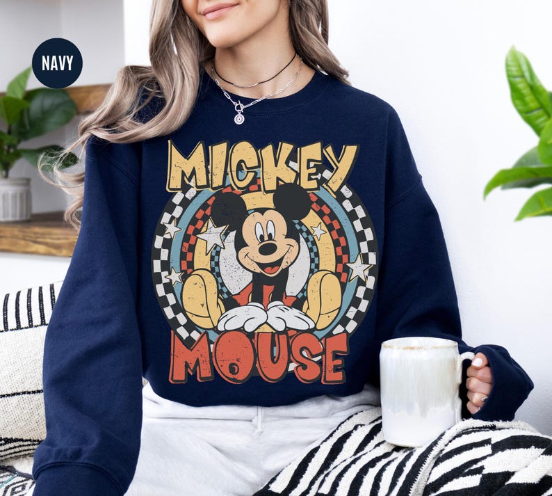 Puede incluir: Sudadera azul marino con un dise&ntilde;o de Mickey Mouse de estilo vintage. El dise&ntilde;o incluye a Mickey Mouse, el texto "Mickey Mouse", estrellas y un patr&oacute;n de cuadros. La sudadera tiene cuello redondo y mangas largas. Se sostiene una taza blanca.