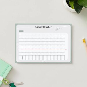 May include: A weight tracker notepad with the German word "Gewichttracker" at the top. The notepad has a light green border and a grid for tracking weight loss. A pencil, a small green notebook, and a plant are also visible.