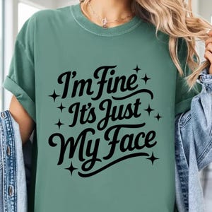 May include: A green t-shirt with the text "I'm Fine It's Just My Face" in black script. The text is surrounded by small star shapes. The shirt has short sleeves and a crew neck.