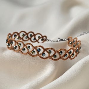May include: A delicate, handmade bracelet featuring a repeating pattern of interconnected loops in a warm brown color. The bracelet is adorned with small, iridescent beads that catch the light, creating a subtle sparkle. The clasp is silver-toned.